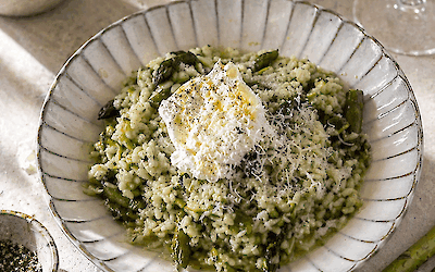 Romige risotto verde met groene asperges, basilicum en citroen, geserveerd met burrata en geraspte kaas op een licht bord, met verse asperges en olijfolie op de achtergrond