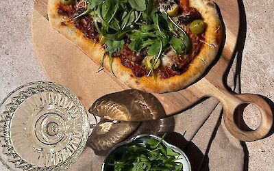 Pinsa with goat’s cheese, rocket, olives and sun-dried tomatoes on a wooden board with a glass of white wine and a bowl of rocket