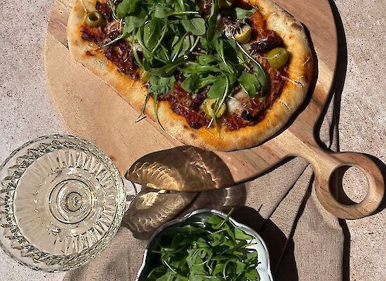 Pinsa with goat’s cheese, rocket, olives and sun-dried tomatoes on a wooden board with a glass of white wine and a bowl of rocket