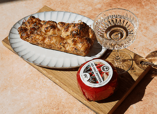 Croissant breekbrood met gesmolten Henri Willig Biologische Koekaas met Chili en hot honey, geserveerd op een houten plank met een glas wijn.