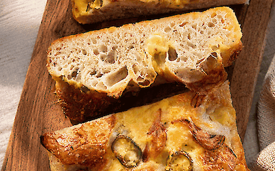 Focaccia au levain garnie de fromage de Jersey bio Henri Willig, d'oignons rouges caramélisés et de piment jalapeño, servie sur une planche en bois.