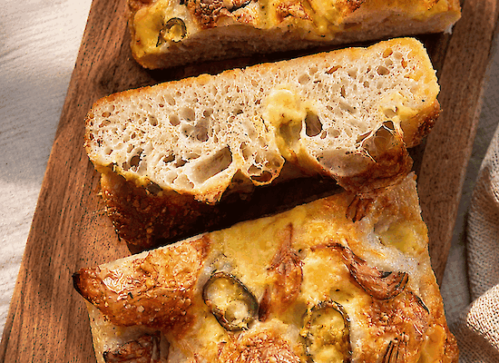 Zuurdesem focaccia met Henri Willig Biologische Jersey Koekaas, gekaramelliseerde rode ui en jalapeño, op een houten serveerplank.
